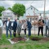 People in business suits hold place shovels in piles of dirt near a small flowering tree.