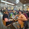 Students with full plates of pancakes, sausage, ice cream and donuts sit at a table in a full cafeteria and smile at the camera.