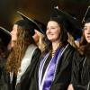 Graduates in caps and gowns stand in an auditorium, smiling and looking toward a stage.