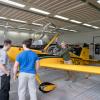 People look on as a pilot opens the canopy of a small yellow airplane.