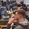 Students work at a long row of desks outfitted with computers and electronics equipment.