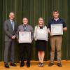 Four people stand on a stage. Three are holding framed certificates.