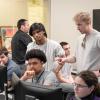 Students sit behind computer screens while two others stand behind them, one pointing toward a screen.