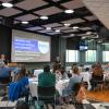 In a conference room, adults sit around round tables and look toward a speaker behind a lectern. A large projection screen shows the title of a session: "Breaking Silos Through Collaboration."