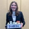 A high school student in a business suit holds a box that says "DiaBEDic" and has compartments with lights that hold a variety of snacks and juices.