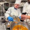 A student in a chef uniform adds ingredients to a large stainless steel bowl filled with liquid while another student in a chef uniform mixes it with an immersion blender.