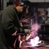 A student in a welding helmet and protective gloves welds a piece, creating light and smoke.