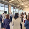  Group of adults standing in a semicircle inside a large industrial-style room while one person speaks; the space has exposed ductwork, fluorescent ceiling lights, concrete surfaces, and large windows showing a cityscape outside.