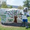The Penn College Wildcat mascot stands next to The Rock, a large boulder on campus that serves as a billboard. The Rock is painted white with a tree that has blue and green hand prints for leaves. The Rock is also painted with the phrases "Donors That Save" and "Donate Life."