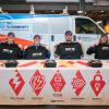 Four Penn College students sit at a table draped in a Tri-M tablecloth. Each is holding a Tri-M T-shirt and wearing a Tri-M hat. Behind them is a Penn College van that has Penn College branding and the words "Connecting Campus to Community: Electrical Technology at Penn College."
