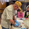 Children watch as an instructor wearing safety glasses and blue gloves uses his hands to mix concrete on a paper plate.