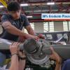 Several students wearing safety glasses work together on an aircraft engine and propeller inside an aviation training hangar.
