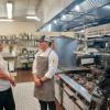 A reporter and a person in a chef uniform talk in a commercial kitchen. A television camera is in the foreground.