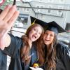 Two students wearing black graduation caps and gowns lean in toward each other and smile widely.