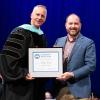Penn College President Micheal J. Reed, wearing graduation regalia, and alumnus Stephen Griegel stand on the Community Arts Center stage holding a framed certificate.