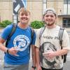 Two student make hand signals with their thumbs and pinkies extended. Penn College's Bardo Gymnasium is visible behind them.