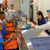 In a cafeteria at the Little League International complex, two adults serve plates of food to children in baseball uniforms.