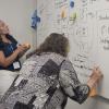 Two educators work side by side at a whiteboard filled with mathematical calculations.