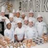A group of students and an instructor, all in chef uniforms, stand behind a counter. The counter is filled with packaged backed goods. The wall behind them is white with a large graphic that says "The Market."