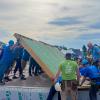 A large group of people, mostly wearing blue T-shirts and hard hats, raises a wall on a foundation.
