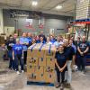 A group of people stand around a palate of cardboard boxes in a warehouse.