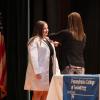 A woman adds a pin to a student's white medical coat. They are standing on a stage.