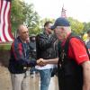 A man wearing a United States Navy vest and red shirt that says "veteran" on the sleeve shakes hands with people lining a sidewalk that leads from a bus to Penn College's Bush Campus Center.