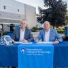 Penn College President Michael J. Reed and Dave Lambert, group president of ITW welding, sit at a table outside of Penn College's Lycoming Engines Metal Trades Center with paperwork in front of them and pens in their hands. Miller welding equipment is positioned behind the table. On the table are a Miller welding helmet and a Pennsylvania College of Technology banner. 